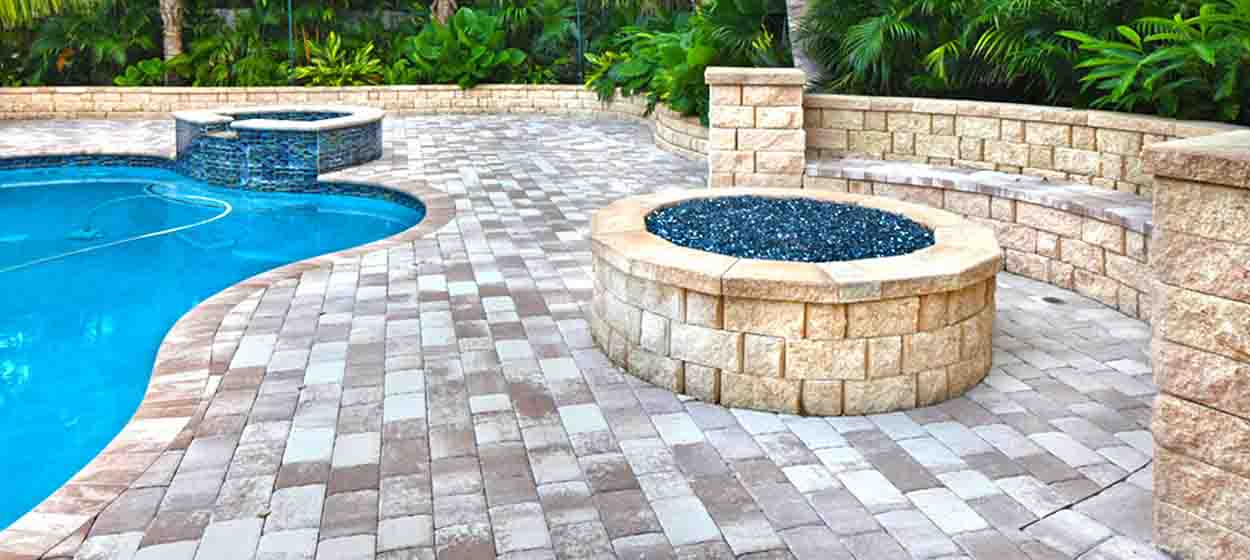 Travertine-toned paver pool deck with stone fire pit and seat wall by Pavers Depot